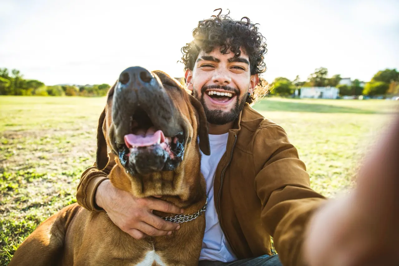 A smiling dog walker outdoors in a park beside a happy brown hound