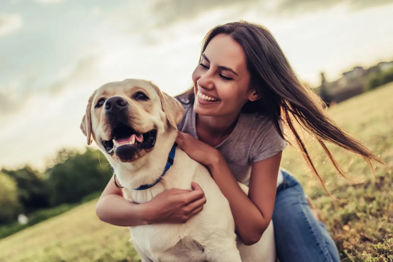 A woman crouched in a park with her arms around a happy yellow Labrador